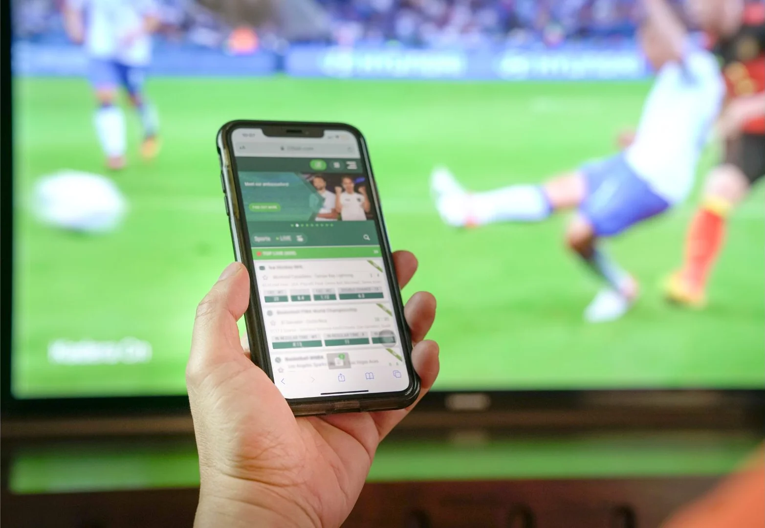 Mão segura celular com aplicativo de apostas diante de uma TV que transmite um jogo de futebol